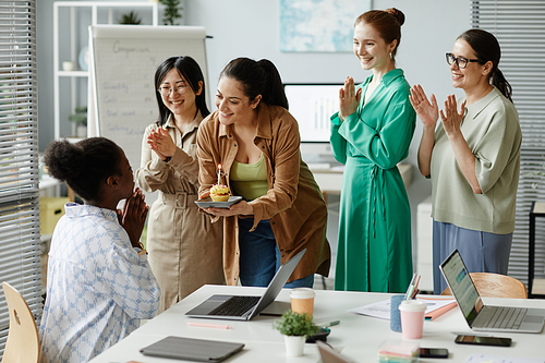 Group of businesswomen clapping hands and congratulating their colleague at office with cupcake with candle