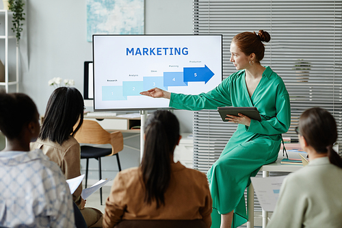 Young businesswoman pointing at screen of monitor with marketing graphs while explaining new strategy to colleagues at training