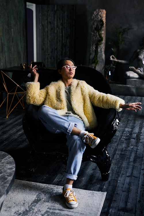 Vertical full length portrait of Asian man in extravagant outfit lounging in leather armchair at dark studio