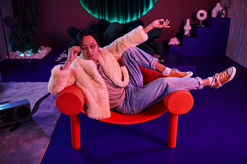 Full length portrait of Asian man wearing extravagant outfit lying across chair in pink neon light