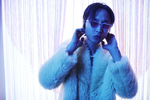 Waist up portrait of young Asian man wearing sunglasses in nightclub lit by blue neon light, copy space