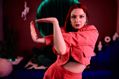 Waist up portrait of young woman dancing vogue style in pink neon light with focus on hand movements and looking at camera