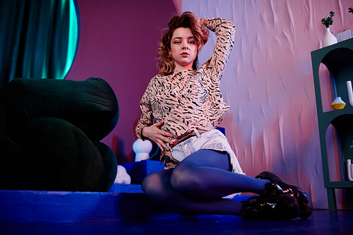 Full length portrait of sensual young woman sitting on floor in club and posing vogue style in neon light, copy space