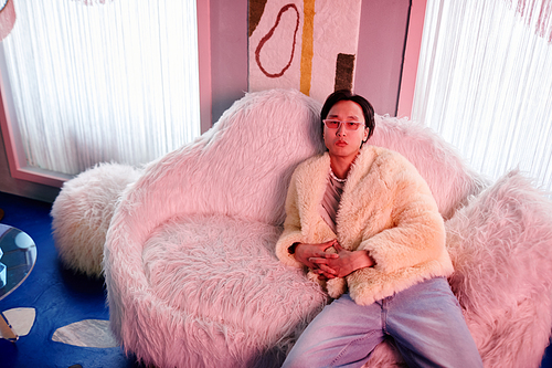 Portrait of Asian young man dressed in fur coat and party outfit relaxing in clu lounge lit by pink neon lights