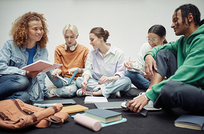 Happy teenage girl with copybook looking at her African American boyfriend during discussion of lecture notes among their classmates