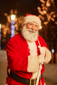 Vertical portrait of traditional Santa Claus holding sack with presents and smiling at camera in night city