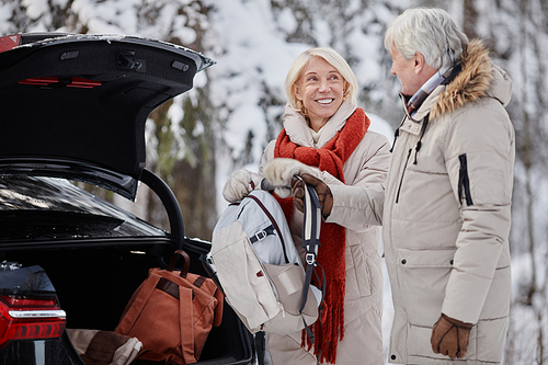 Side view portrait of smiling senior couple opening car trunk while enjoying winter getaway in nature together, copy space