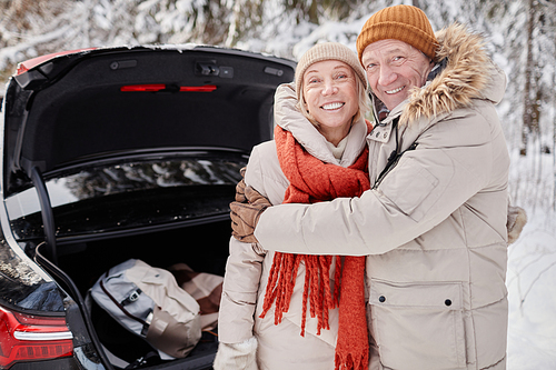 Waist up portrait of smiling mature couple enjoying winter getaway in nature while standing by car trunk and looking at camera, copy space