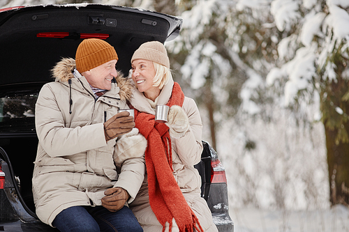 Smiling senior couple enjoying cup of hot coco outdoors in winter while sitting in car trunk, copy space