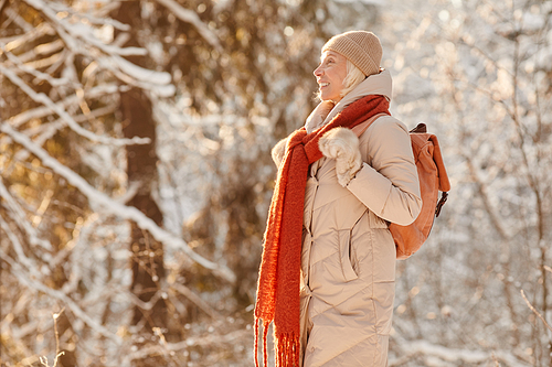 Side view portrait of smiling senior woman enjoying hike in winter forest lit by sunlight, copy space