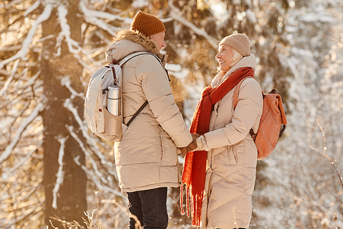 Side view portrait of smiling senior couple enjoying hike in winter forest and holding hands in sunlight, copy space