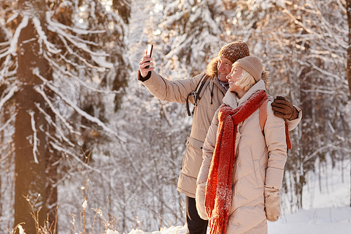 Side view portrait of active mature couple taking selfie photo while enjoying hike in winter forest, copy space