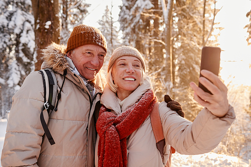 Waist up portrait of happy mature couple taking selfie photo while enjoying hike in winter forest, copy space