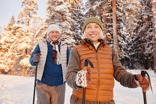 Waist up portrait of active senior couple walking with poles in winter forest and smiling at camera