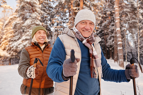 Waist up portrait of happy senior couple walking with poles in winter forest and smiling at camera