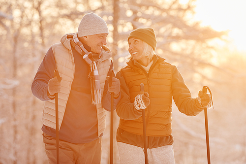 Waist up portrait of active senior couple enjoying Nordic walk with poles in beautiful winter forest lit by sunlight