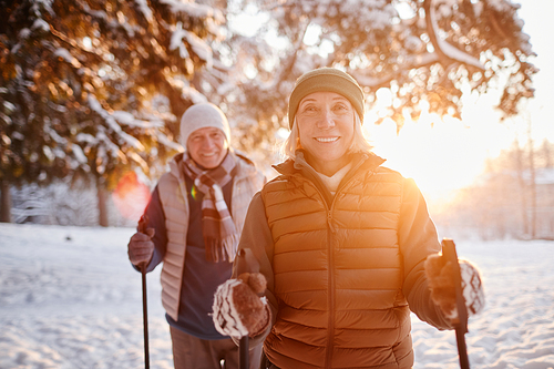 Waist up portrait of smiling mature couple enjoying Nordic walk in winter forest at sunset