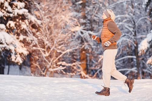 Side view portrait of sporty mature woman running in winter forest lit by sunset light, copy space