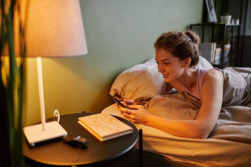 Young woman lying on bed in bedroom and communicating online using her smartphone