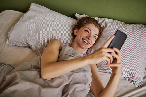 Young happy woman reading message in her smartphone and smiling while resting in her bed during weekends