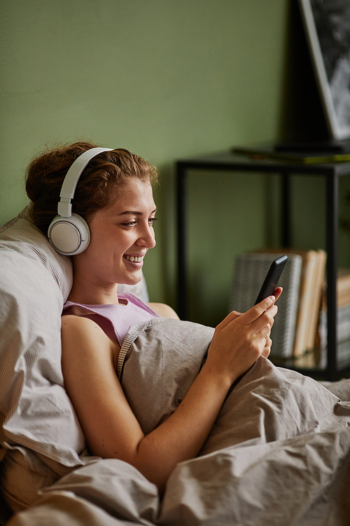 Happy girl in wireless headphones resting in her bed and listening to music on her smartphone