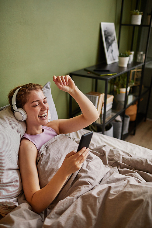 Happy girl in headphones having video call on smartphone with her friend while resting on bed in bedroom