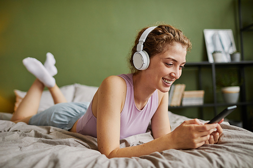 Young woman in wireless headphones resting on her bed and talking online using her smartphone