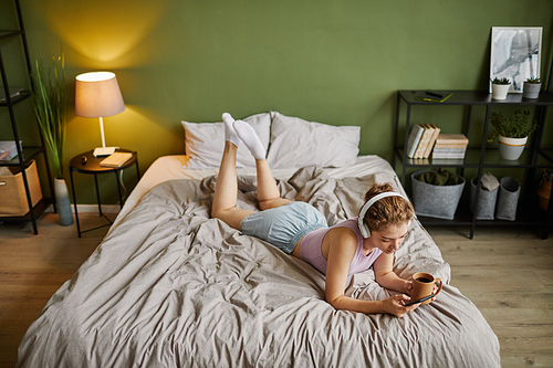 High angle view of young woman in headphones using mobile phone while lying on her bed with cup of coffee