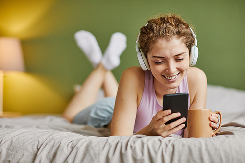 Smiling girl in wireless headphones communicating online using her smartphone and drinking coffee in bed in the morning