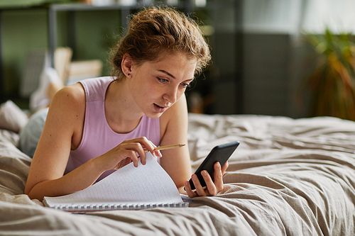 Young girl lying on bed using her smartphone to make notes in notebook