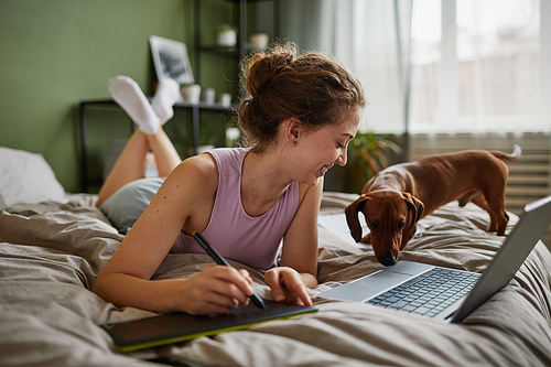 Girl drawing on graphic tablet while lying on bed with laptop and her dog in bedroom at home