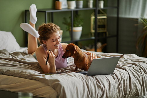 Young girl using laptop and playing with her dog while relaxing in bed during leisure time at home