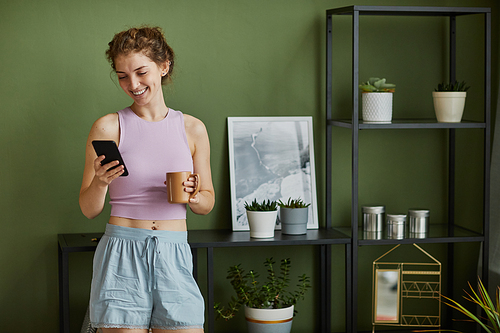 Young happy girl reading message on mobile phone and smiling while drinking coffee in the room