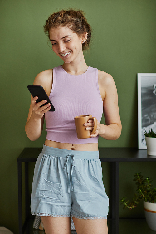 Young happy woman getting message on her smartphone while drinking coffee in the morning in the room