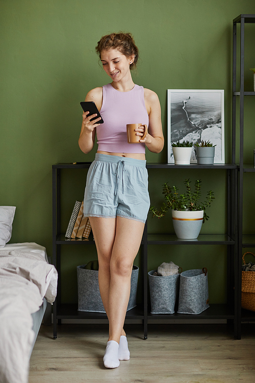 Young woman in domestic clothes communicating online on her smartphone and drinking coffee standing in her bedroom