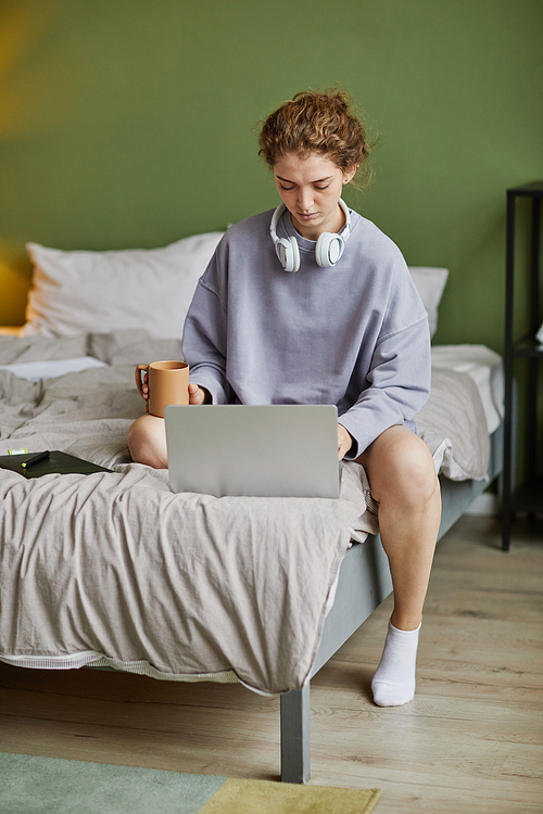 Young woman sitting on bed with cup of coffee and typing on laptop at home