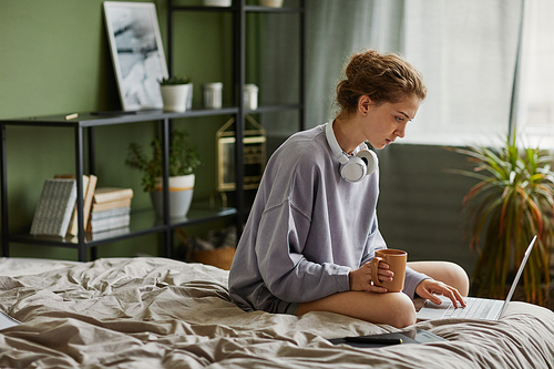 Young woman drinking coffee and using laptop in the morning at home while sitting on her bed