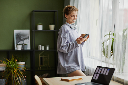 Happy girl using smartphone for online communication, she reading message and smiling while drinking coffee in the morning