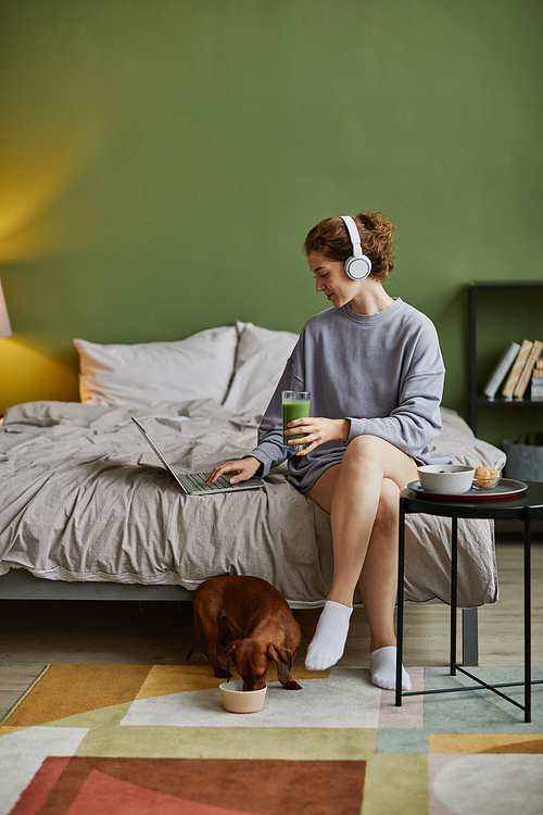 Young woman in headphones communicating online on laptop during breakfast in bedroom