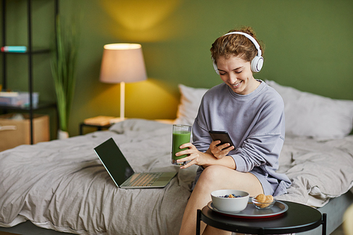 Young woman in wireless headphones drinking smoothie and using mobile phone during breakfast in bedroom