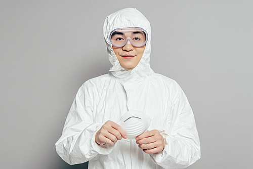 asian epidemiologist in hazmat suit holding respirator mask while  on grey background