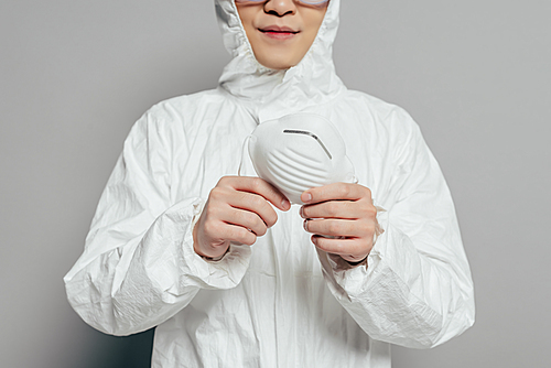 cropped view of epidemiologist in hazmat suit holding respirator mask on grey background