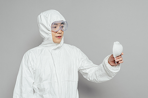 asian epidemiologist in hazmat suit holding respirator mask on grey background