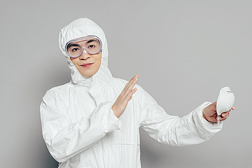 asian epidemiologist in hazmat suit showing no gesture while holding respirator mask and  on grey background