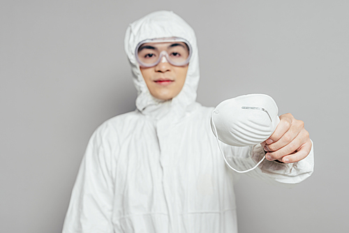asian epidemiologist in hazmat suit showing respirator mask while  on grey background