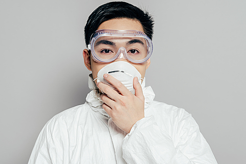 asian epidemiologist in hazmat suit touching respirator mask while  isolated on grey