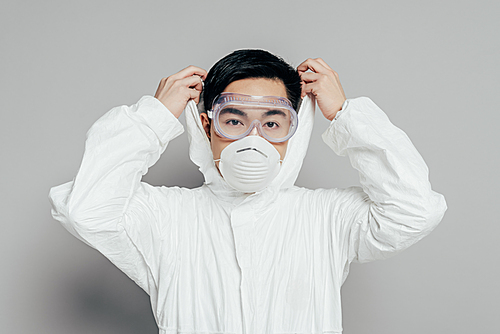 asian epidemiologist in hazmat suit and respirator mask putting hood on while  on grey background