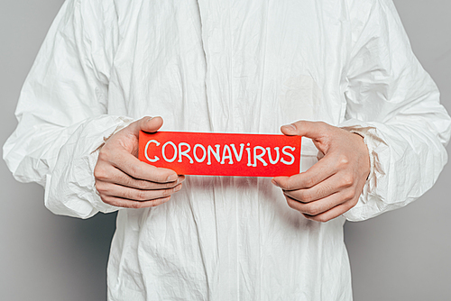 cropped view of epidemiologist in hazmat suit holding warning card with coronavirus inscription on grey background