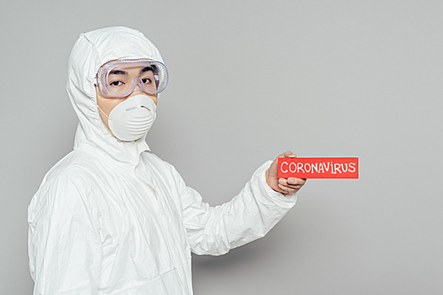asian epidemiologist in hazmat suit and respirator mask holding warning card with coronavirus inscription on grey background