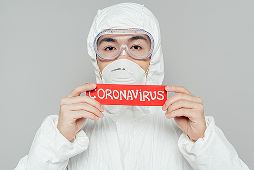 asian epidemiologist in hazmat suit and respirator mask holding warning card with coronavirus inscription isolated on grey